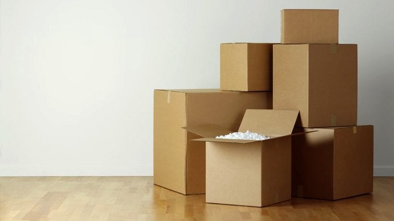 Cardboard moving boxes stacked on a wooden floor, with one open and styrofoam packaging visible.