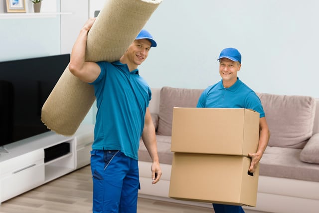 Two professional movers, one holding two boxes and another carrying a rug, smiling and preparing to move out